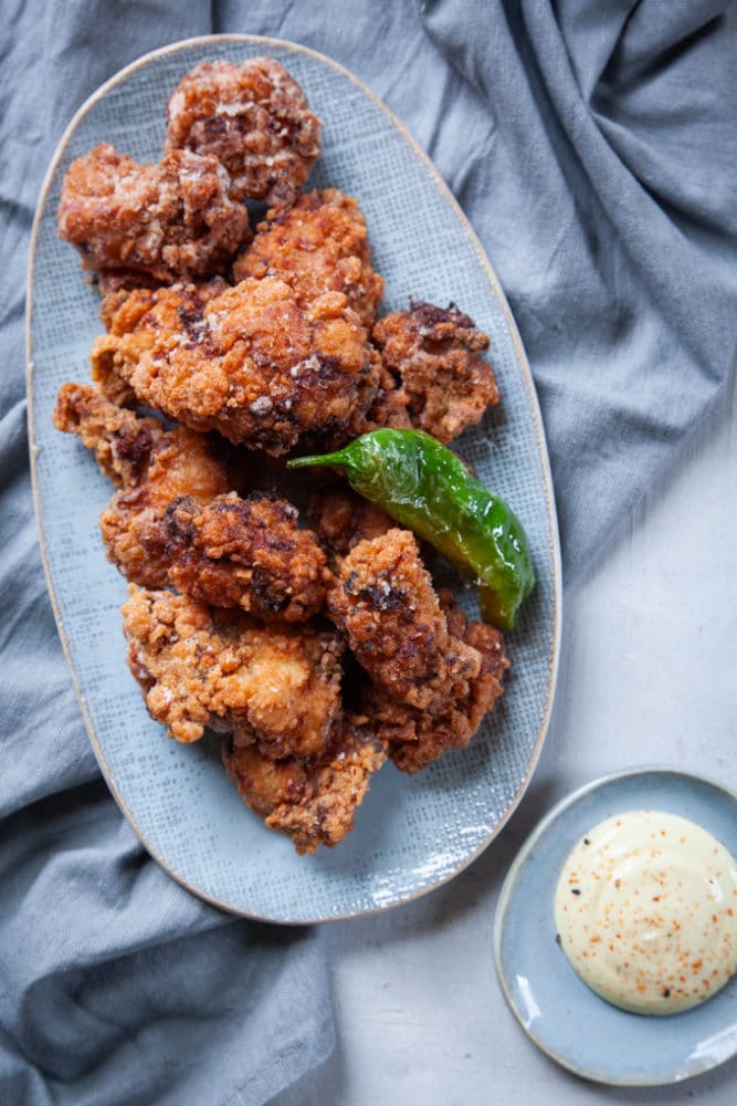 Karaage chicken stacked in a pile on a plate garnished with a blisted shishito pepper and a small plate of Kewpie mayonnaise.