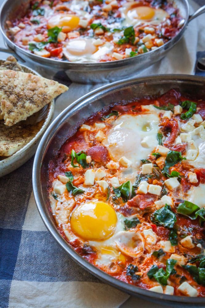 two pans with Shakshuka, a thick tomato sauce dish with eggs cooked directly in it, sitting on a table with a bowl of toasted pita on the side.