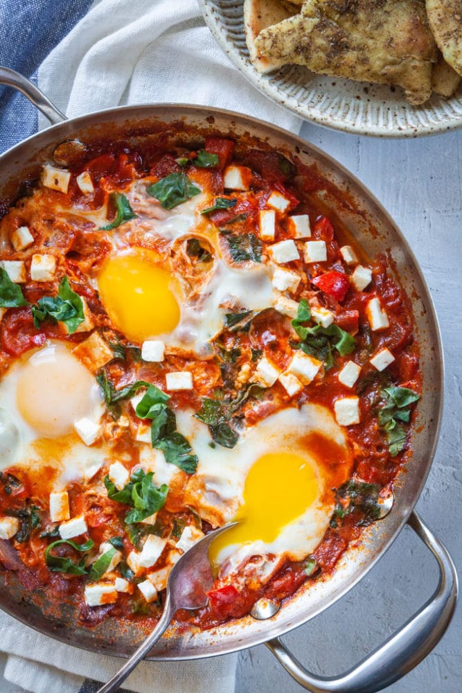 Shakshuka, a thick tomato sauce dish with eggs cooked directly in it, sitting on a table with a bowl of toasted pita on the side.