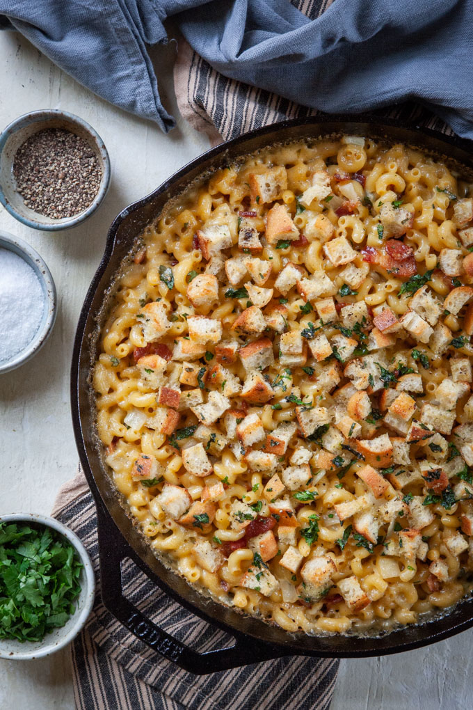 Baked mac and cheese with bacon in a cast iron skillet, surrounded by small bowls of prep ingredients, like salt, pepper and chopped parsley.