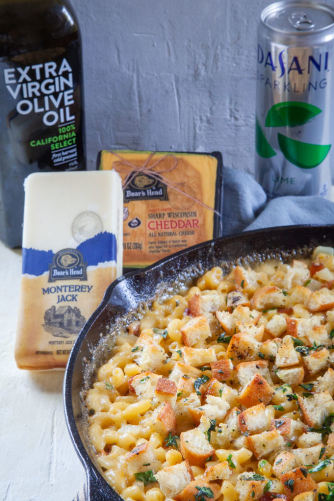 bacon mac and cheese in a cast iron skillet, with Boar's Head cheese, Cobram Estate Extra Virgin Olive Oil and Dasani Sparkling Water behind it.