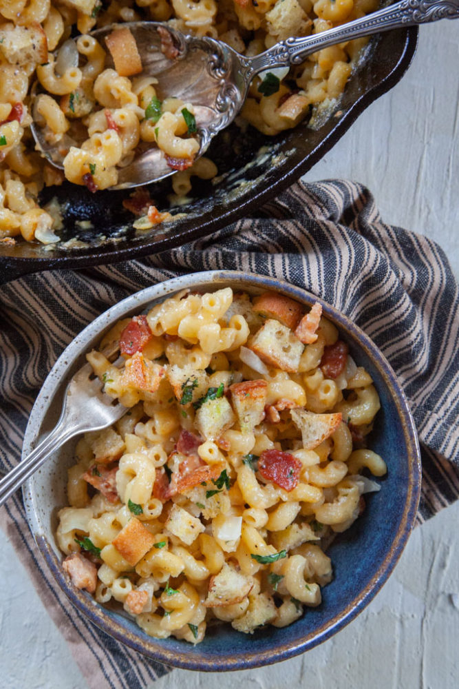 a bowl of homemade mac and cheese with bacon next to a cast iron skillet filled with more mac and cheese. 