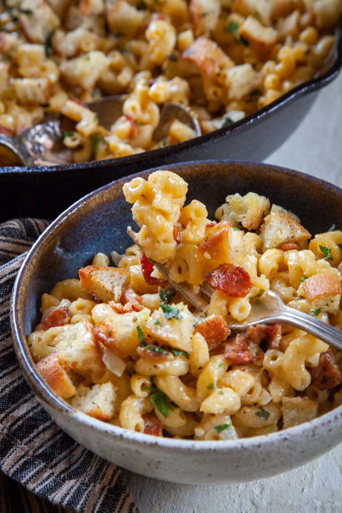 a fork filled with homemade baked mac and cheese with bacon over a bowl of the mac and cheese and a skillet in the background, filled with the bacon mac and cheese.