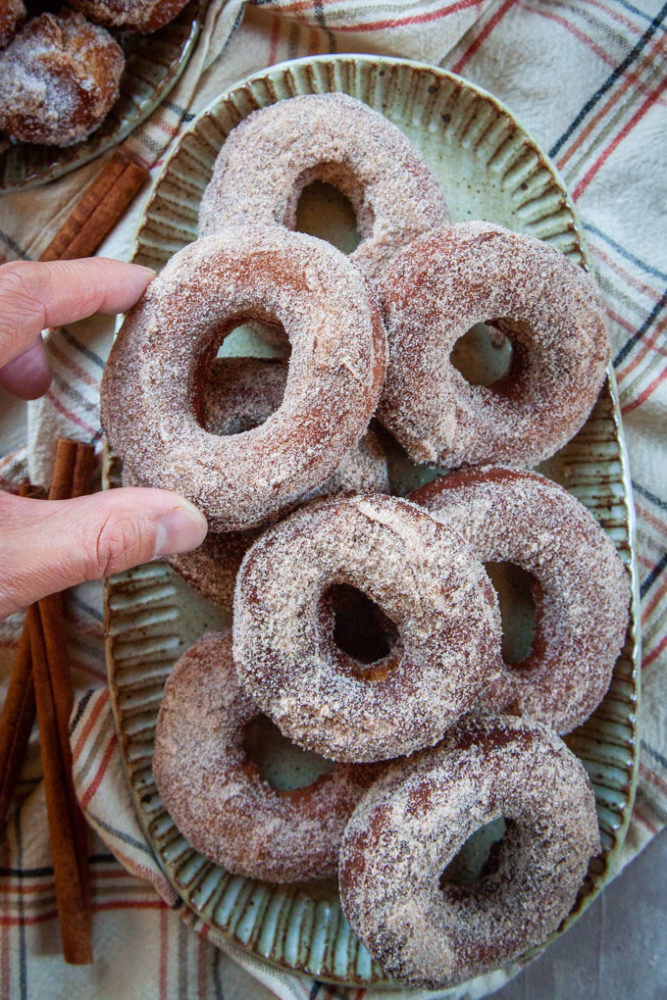 a hand reaching for an apple cider donut on a plate piled with cider donuts.