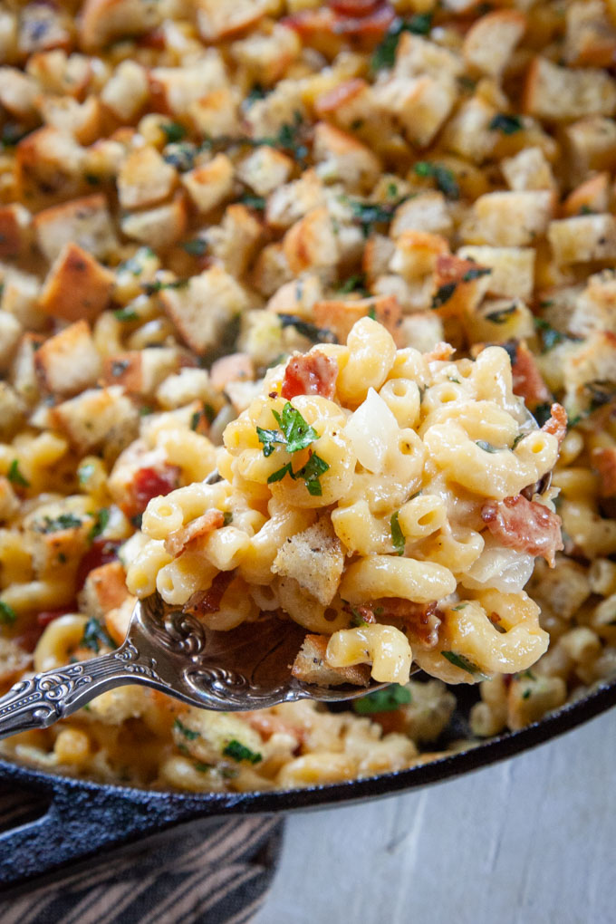 a serving spoon filled with homemade mac and cheese with bacon over a skillet filled with more mac and cheese.