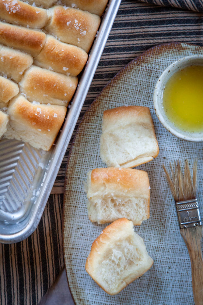Parker House dinner rolls on a serving plate with a brush and small bowl of melted butter.