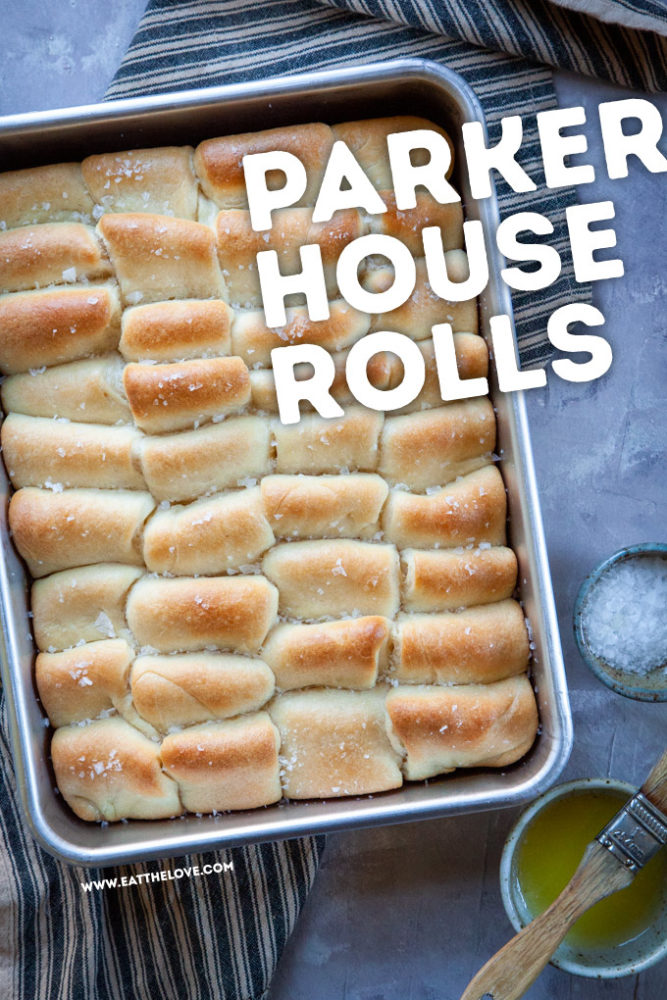 Fresh baked Parker House rolls in a pan sitting on a table with melted butter and flaky salt in small bowls next to baking pan.