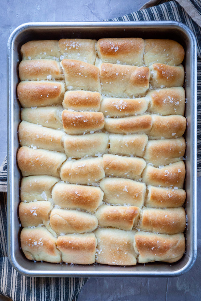 Parker House Rolls baked in a baking pan, sitting on a kitchen towel on a table.