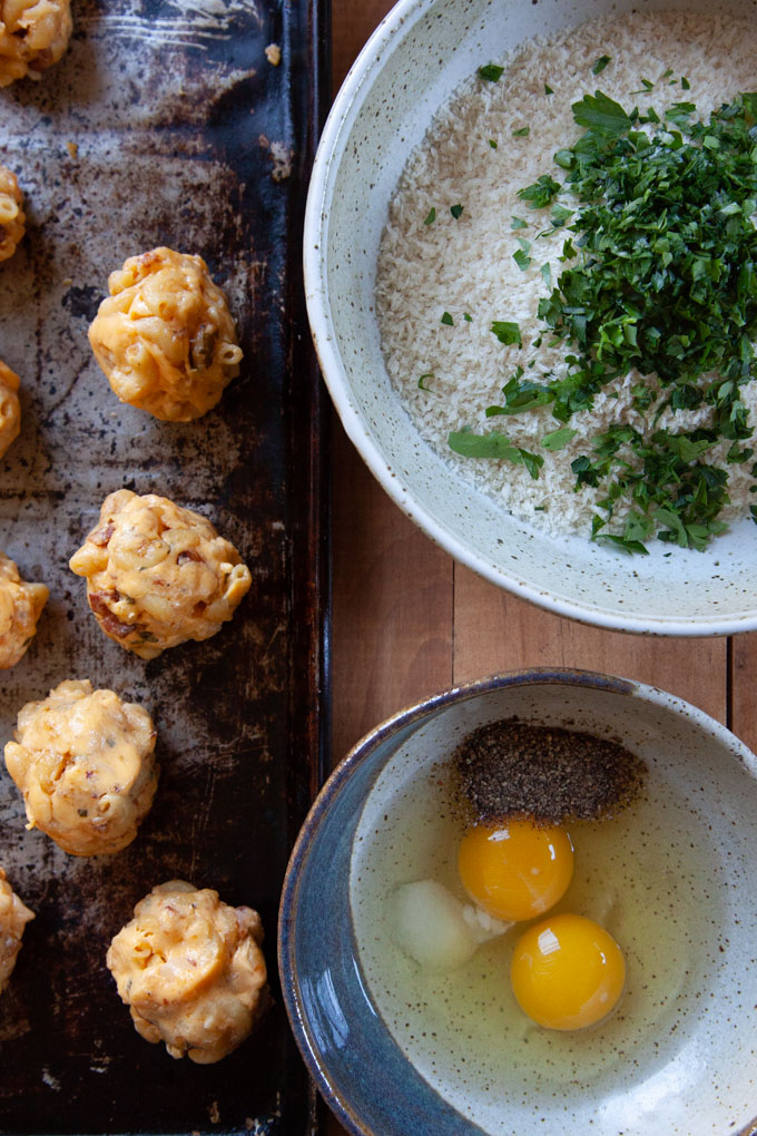 Prepping fried mac and cheese bites with plain mac and cheese balls on a baking sheet next to a bowl with eggs in it and a bowl with panko bread crumbs in it.