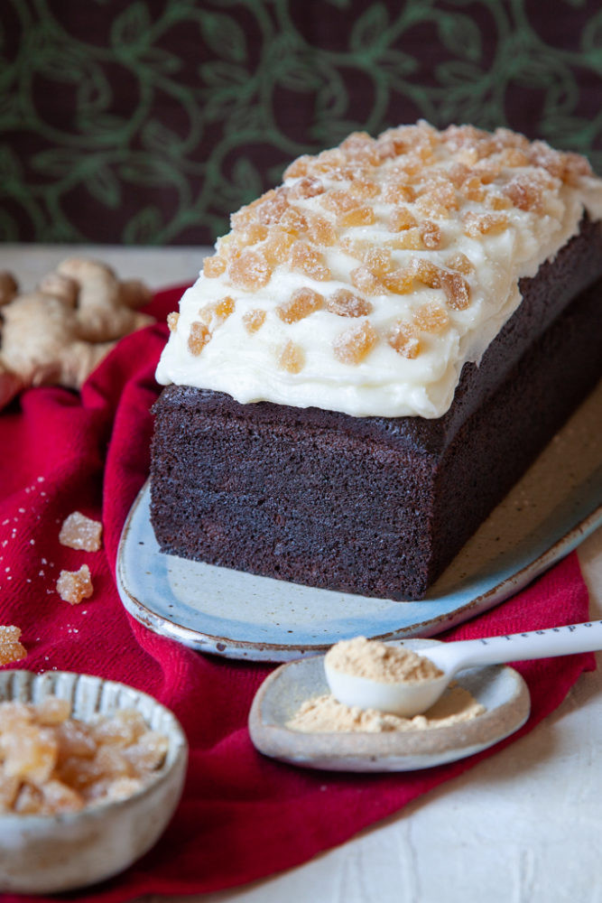 Sliced gingerbread stout cake on a plate surrounded by fresh ginger root, candied ginger, and ground ginger.