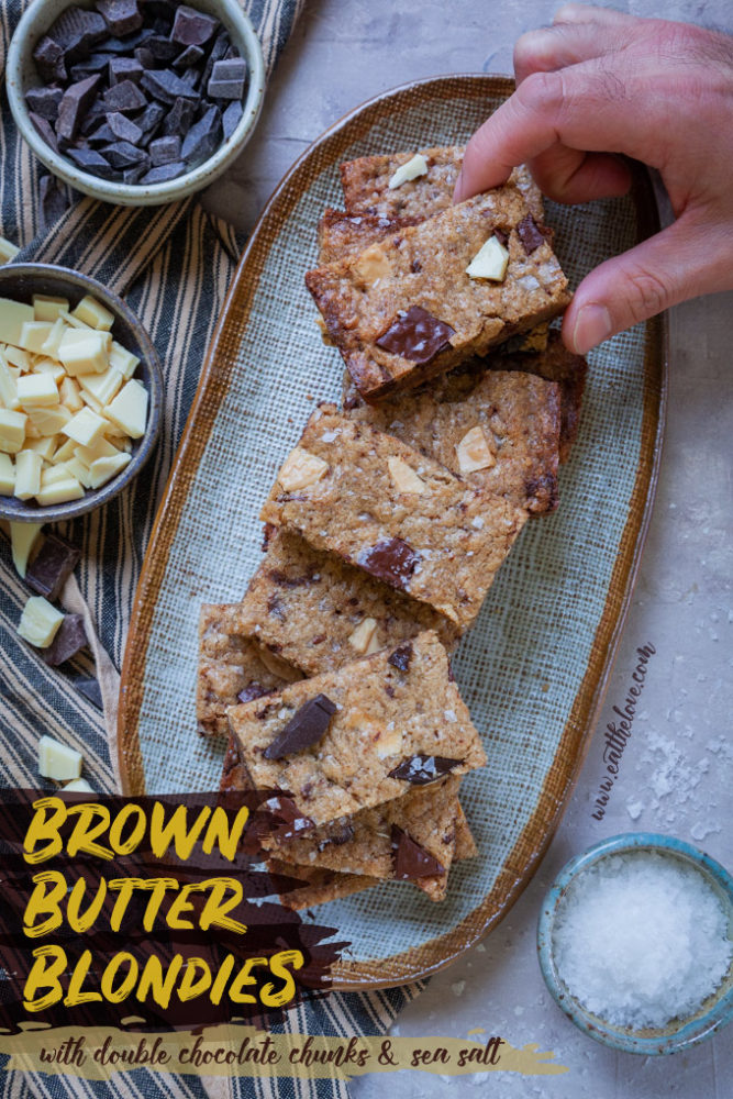 A hand reaching out to grab a brown butter blondie from a plate of blondies, surrounded by chopped chocolate.