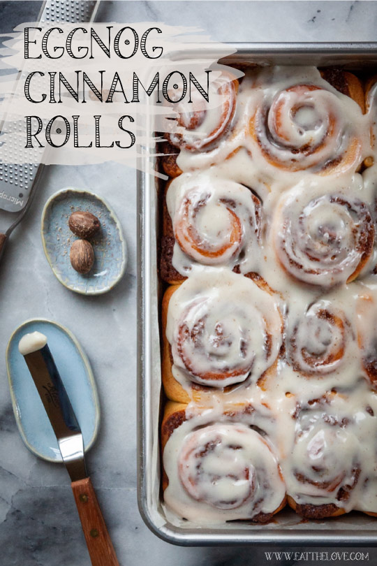 Eggnog Cinnamon Rolls in a pan, next to nutmeg and an offset spatula used to spread the bourbon cream cheese glaze.