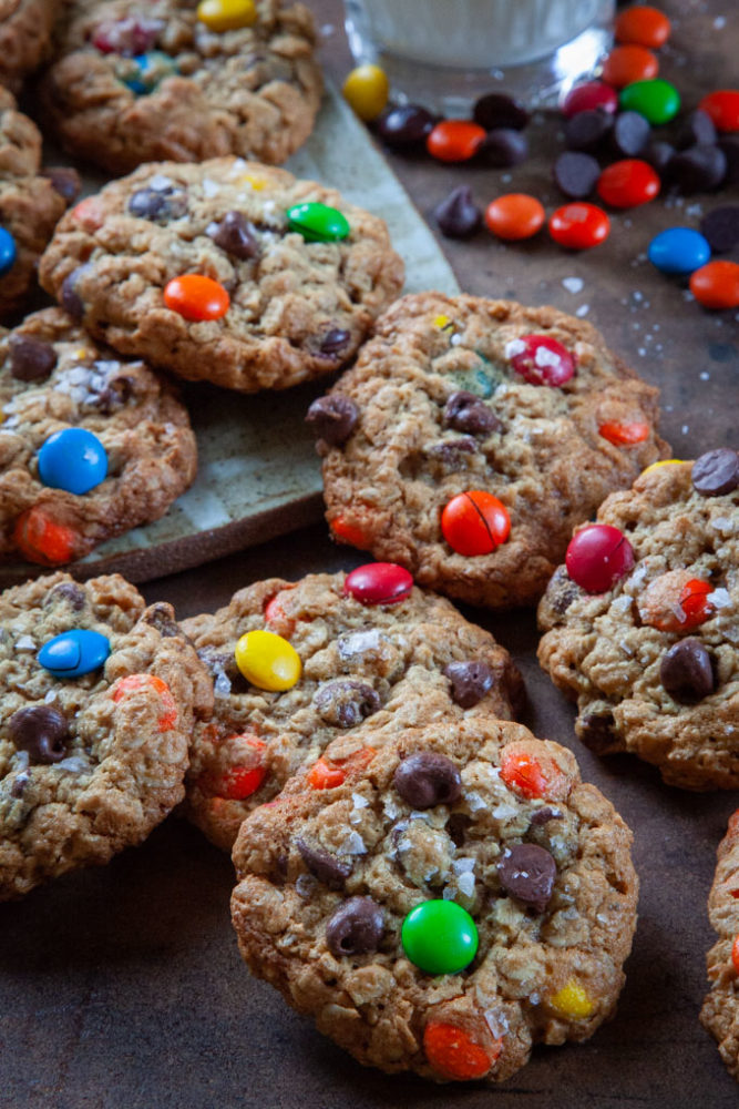 A pile of monster cookies on a table with chocolate chips, m&ms and reese's pieces in the background.