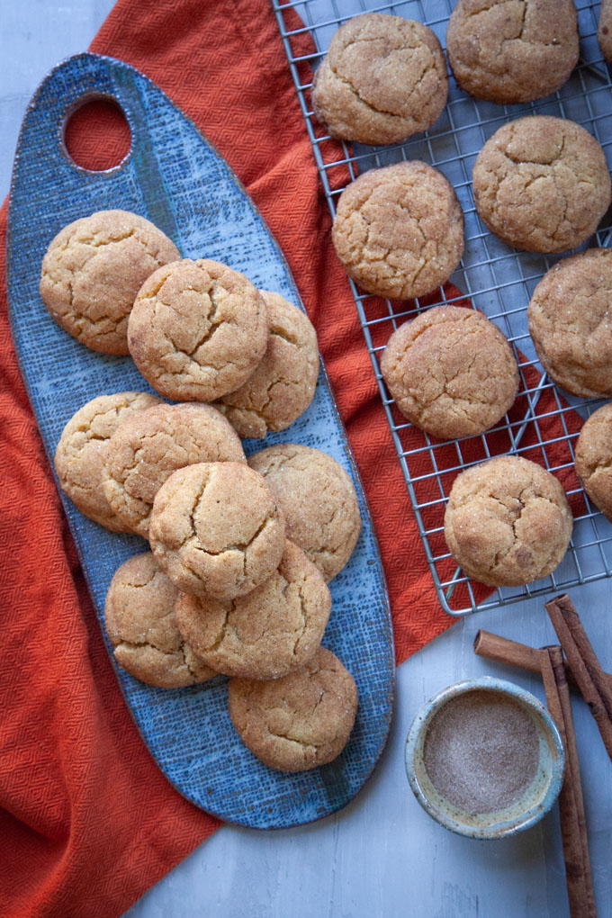 Pumpkin Snickerdoodles on a wire rack and platter next to cinnamon sticks and a small bowl of pumpkin spice blend.