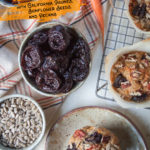 A carrot muffin with prunes, sunflower seeds and pecans on a plate with bowls of ingredients around it.