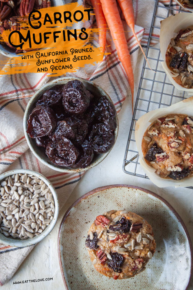 A carrot muffin with prunes, sunflower seeds and pecans on a plate with bowls of ingredients around it.