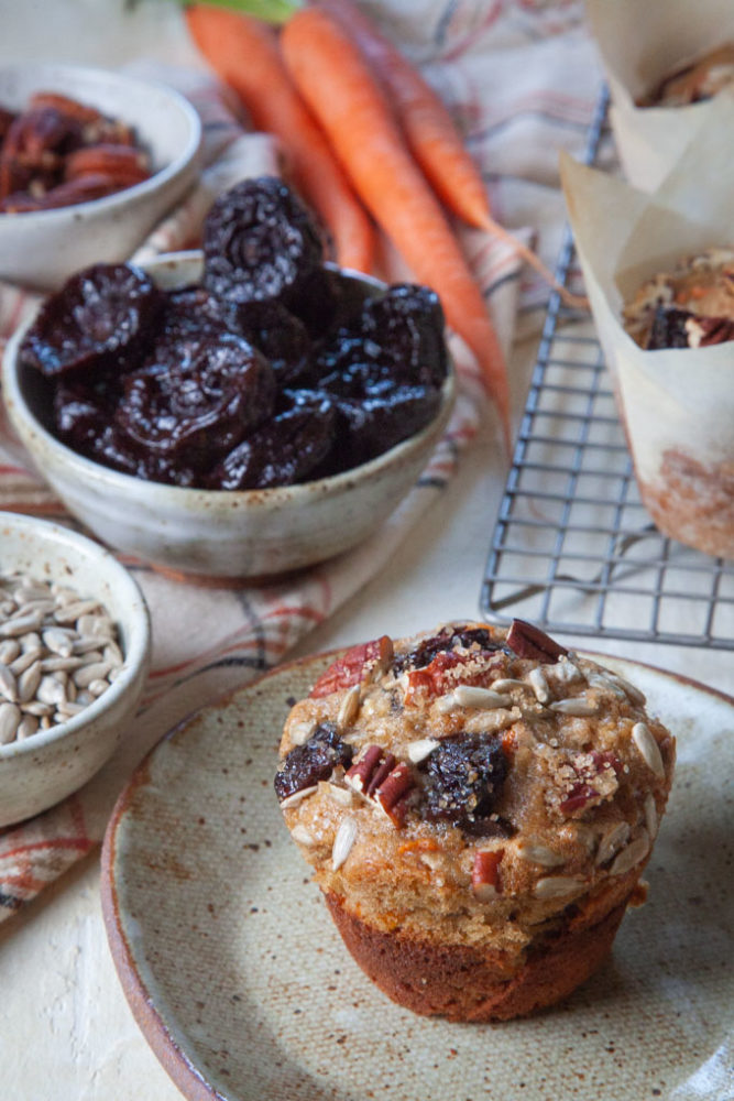 a carrot muffin with prunes, sunflower seeds and pecans on a plate, with bowls of ingredients around the plate.
