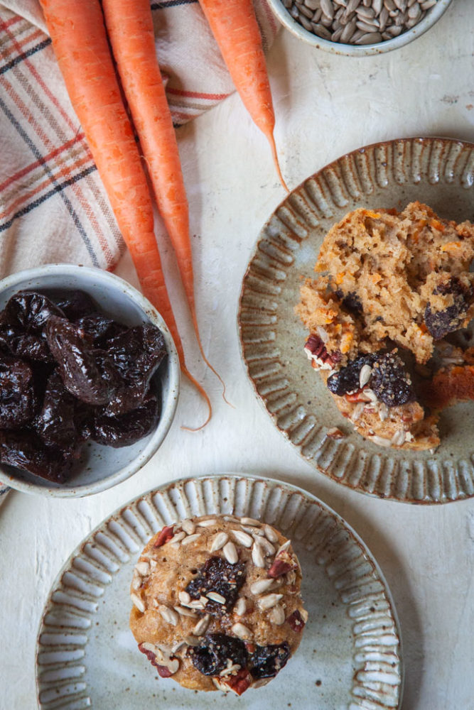 overhead photo of carrot muffins on a plates, with a bowl of prunes and some carrots next to them.