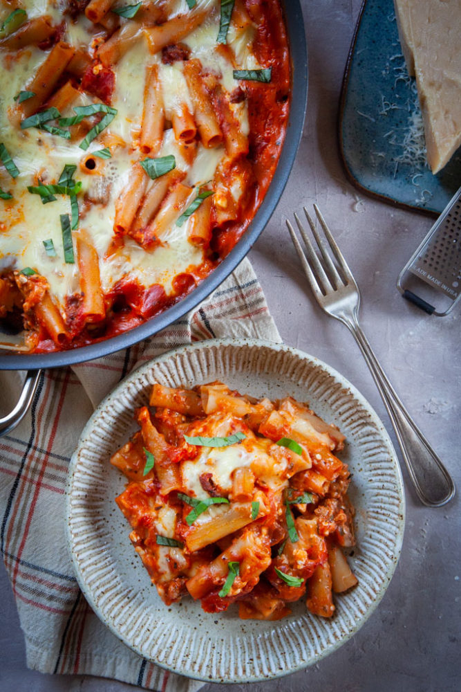 An easy baked ziti recipe in bowl, with the whole skillet of it in next to the bowl.