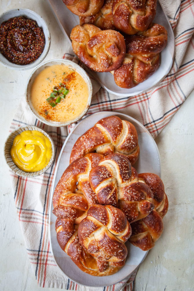 Two plates filled with homemade soft beer pretzels next bowls with a variety of dips like mustards and beer cheese dip.