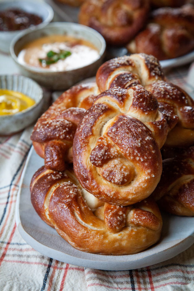 Homemade beer pretzels on a plate, with various dips like mustard and beer cheese dip next to the plate.