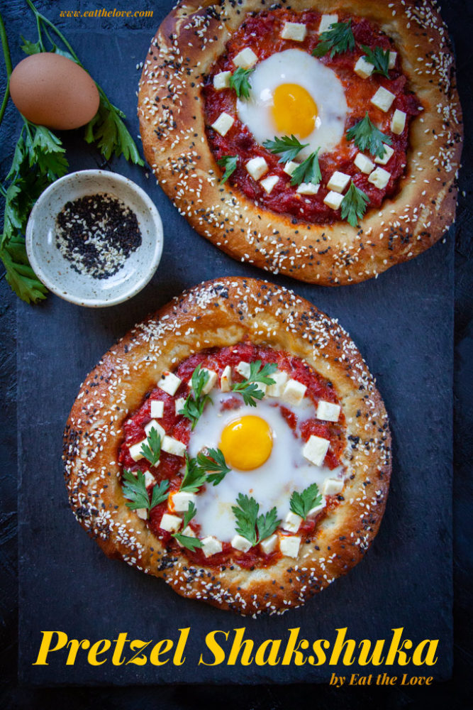 Two pretzel shakshuka sitting on a black slate surface with some ingredients next to it.