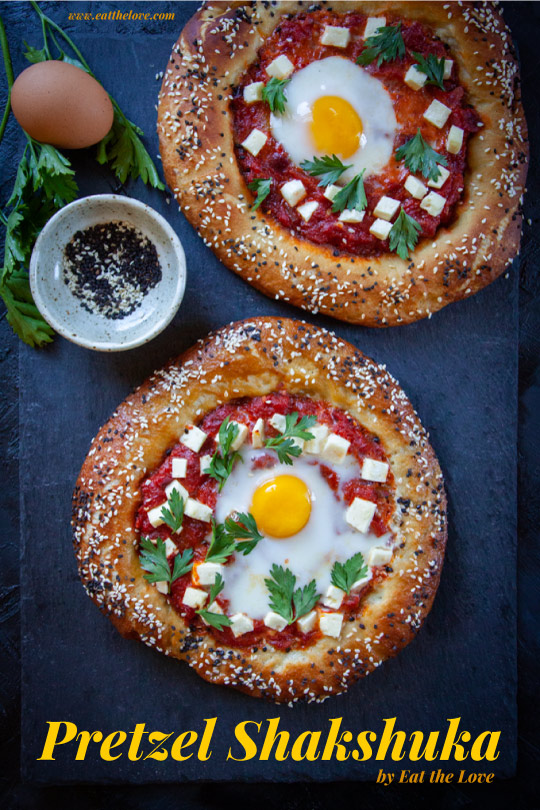 Two pretzel shakshuka sitting on a black slate surface with some ingredients next to it.