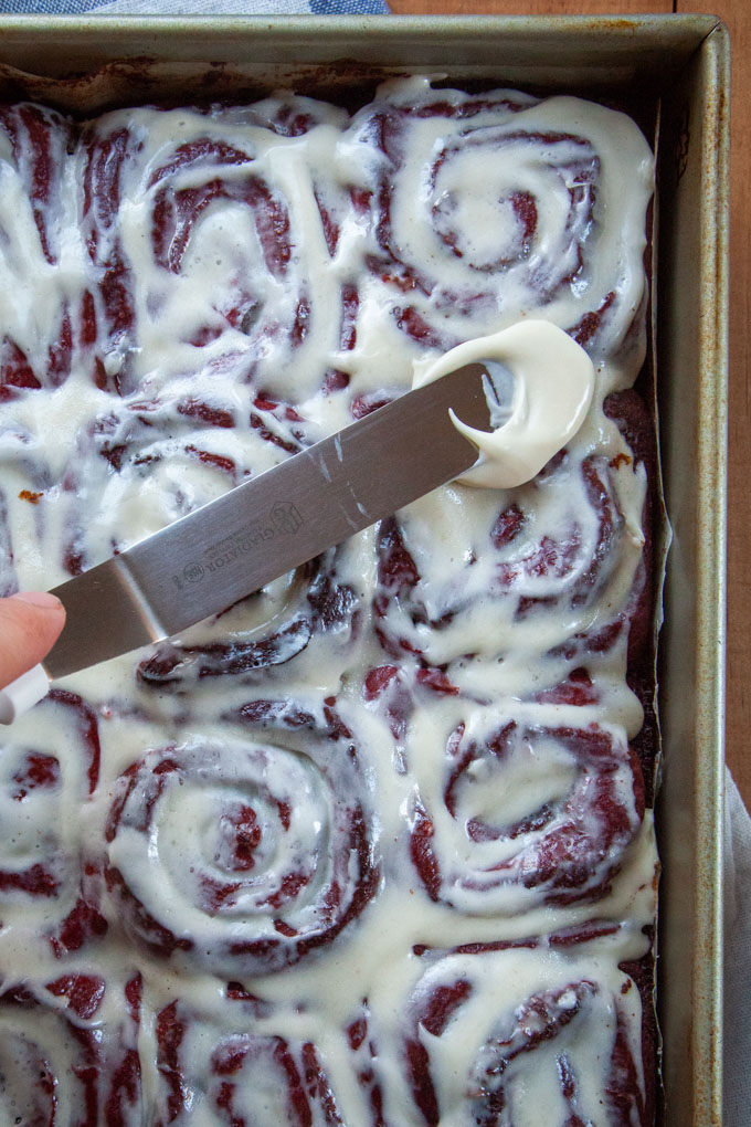 Spreading more cream cheese frosting over the red velvet cinnamon rolls.