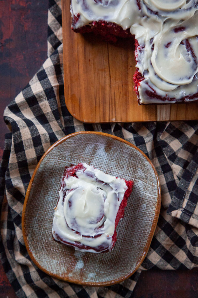 a red velvet cinnamon roll on a plate next to a cutting board with the remaining cinnamon rolls.