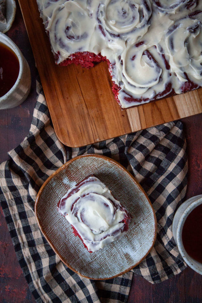 a single red velvet cinnamon bun on a plate, next to the remaining cinnamon rolls and a couple mugs of tea.