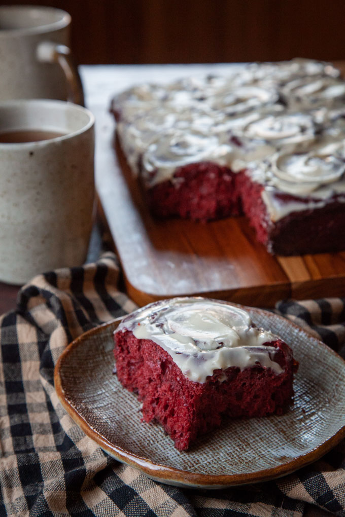 A red velvet cinnamon roll on a plate, with the remaining cinnamon rolls behind it, along with two mugs of tea.