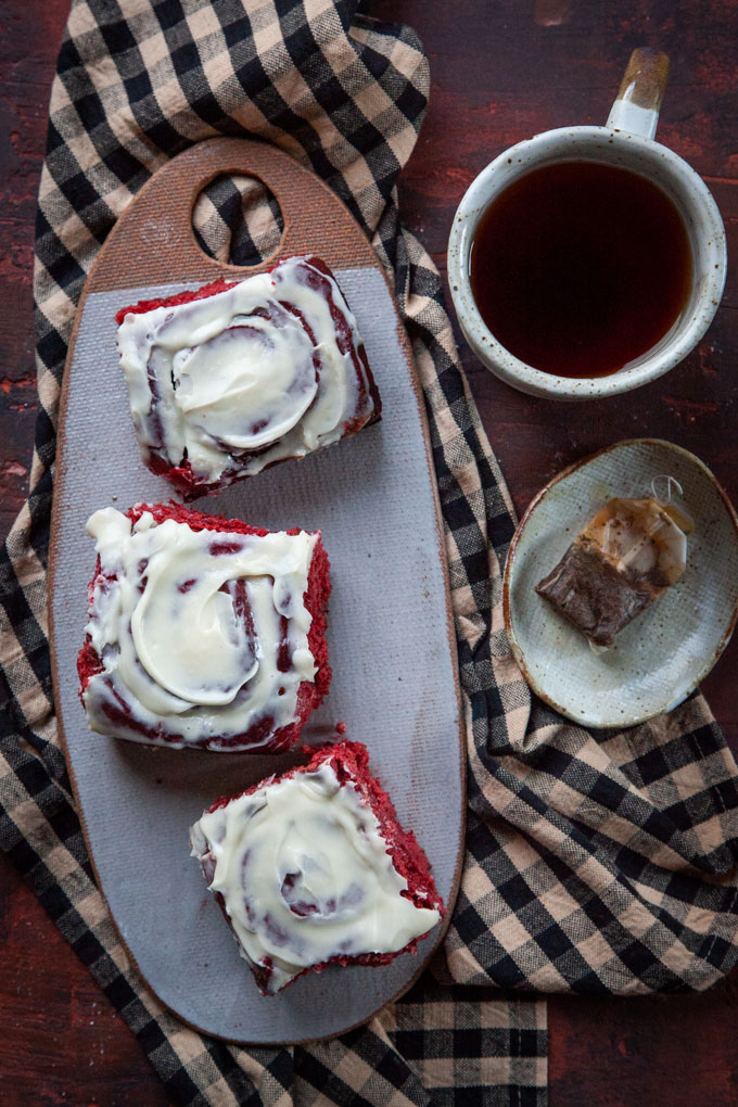 Three red velvet cinnamon rolls on a ceramic cheeseboard next to a mug of tea.