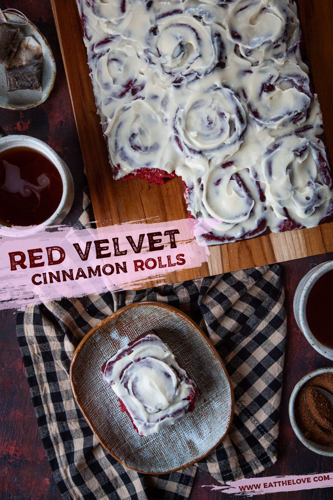 A single red velvet cinnamon rolls on a plate, with the remaining cinnamon rolls on a cutting board, surrounded by two mugs of tea.