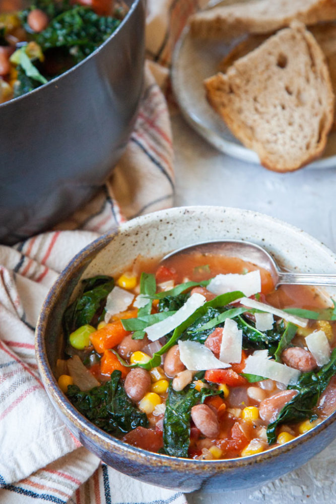 a bowl of minestrone soup next to a large pot of soup and a plate of bread.