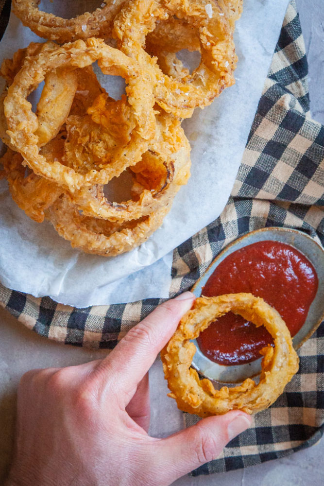 a hand dipping an onion ring in ketchup, next to a plate of onion rings.