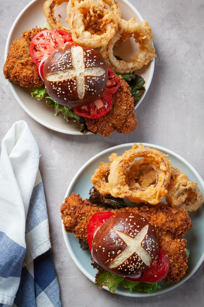 two plates with breaded pork tenderloin sandwiches and onion rings on them.