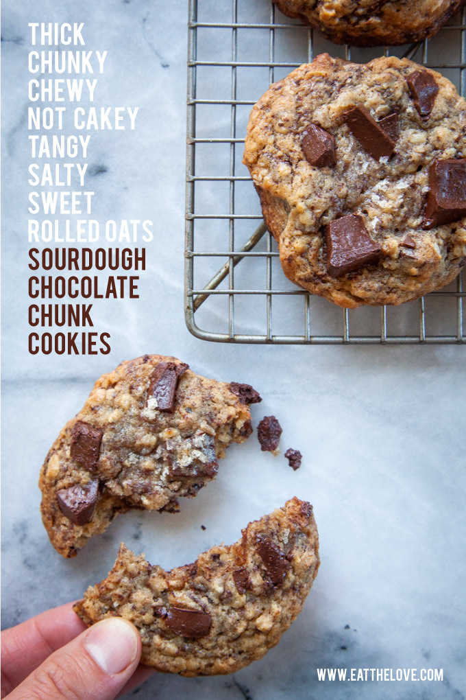 A sourdough chocolate chunk cookie broken in half with a hand reaching out to grab it, with a couple of cookies on a wire cooling rack.