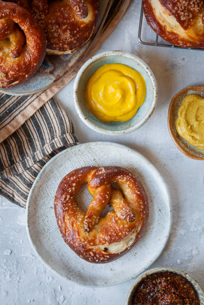 a single sourdough pretzel on a plate with bowls of mustard next to it.