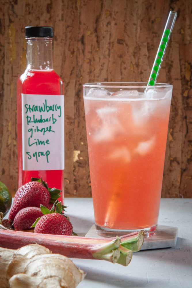 a glass of strawberry rhubarb soda with a bottle of homemade syrup and the ingredients next to it.