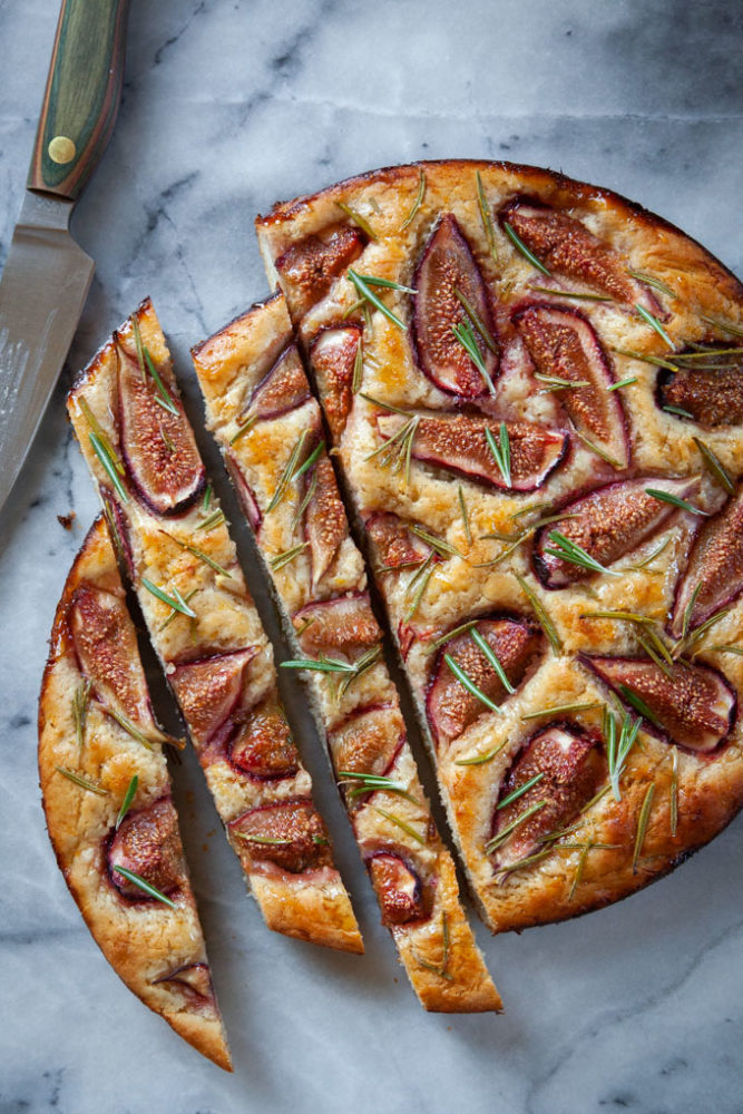 fig rosemary focaccia on a marble surface surrounded by ingredients for the dish.