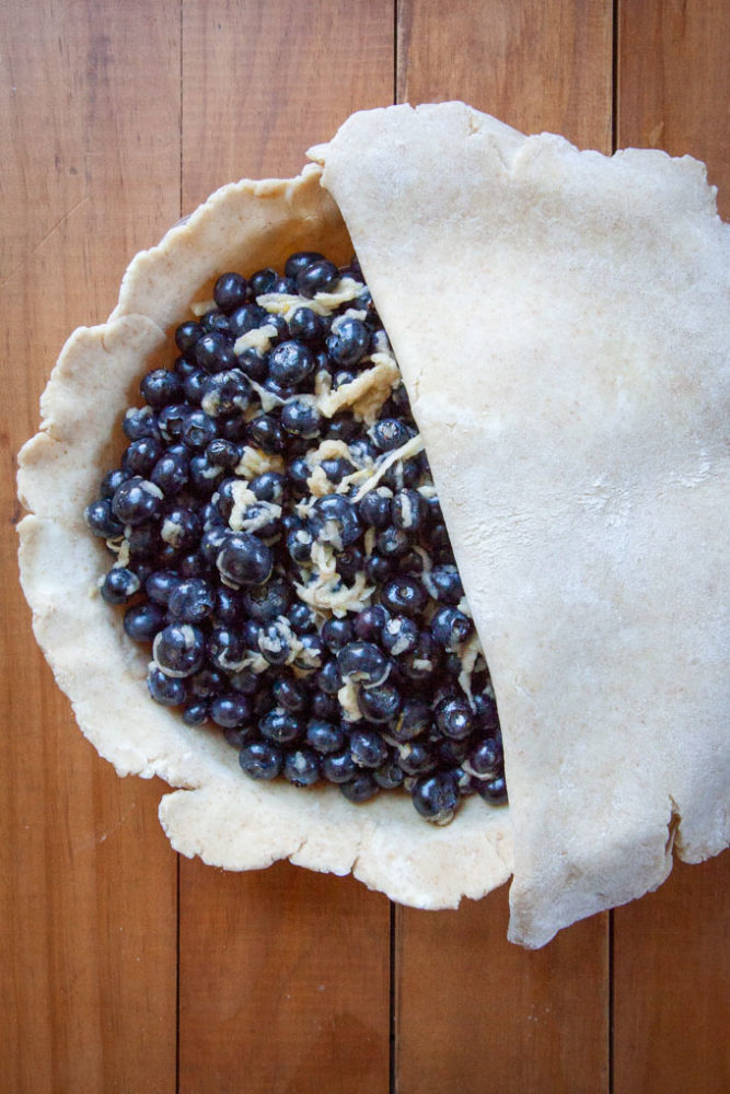 a pie crust filled with blueberry pie filling, being covered with another layer of pie crust.