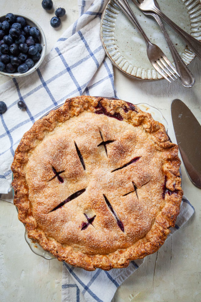 A whole blueberry pie on a table next to some dessert plates.