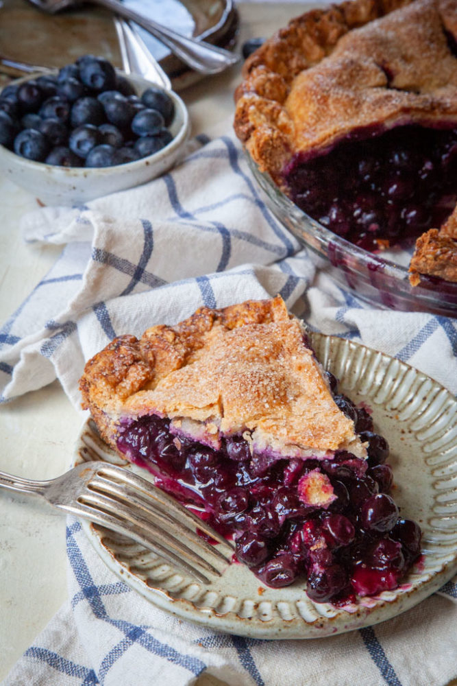 a slice of blueberry pie on a plate, next to the remaining blueberry pie.