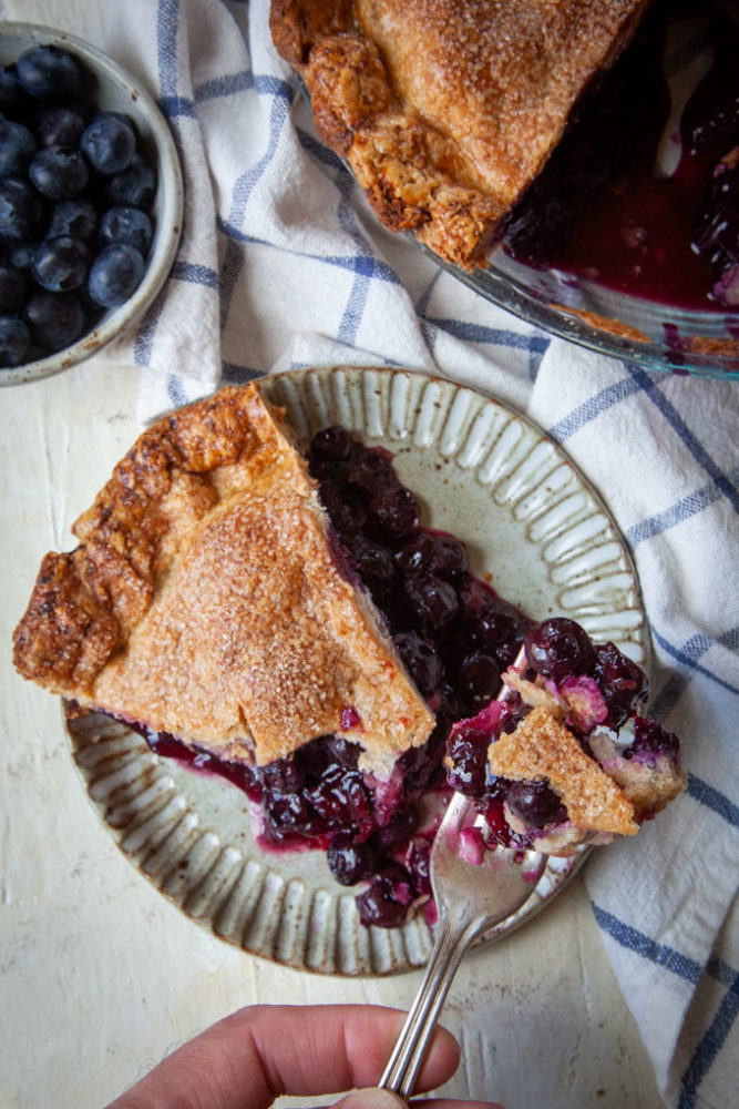 A hand cutting a bite of blueberry pie out of a slice on a ceramic plate.