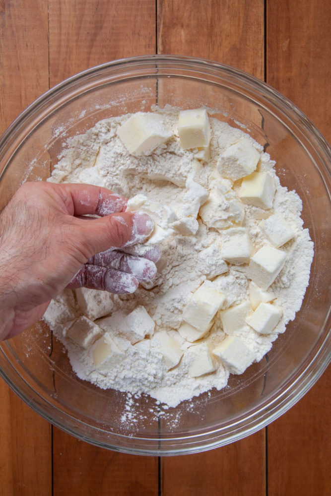 A hand smashing butter in bowl filled with flour to make pie crust.
