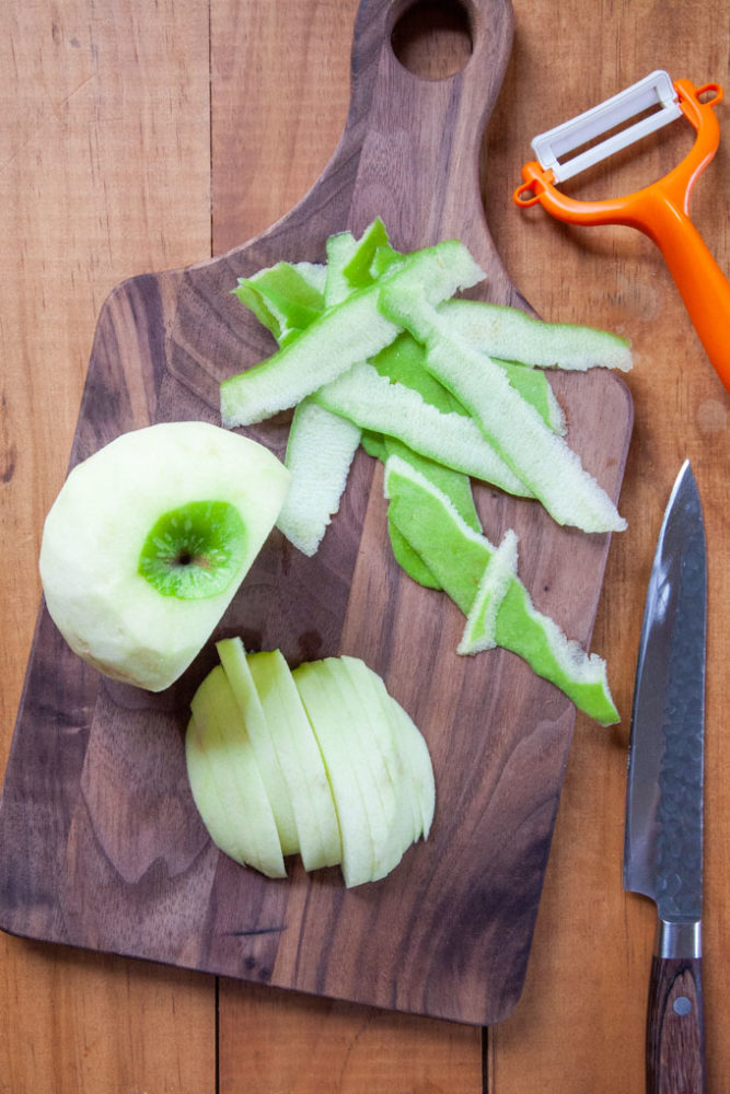 A peeled Granny Smith apple that has been partially sliced on a cutting board with a knife and peeler next to it.