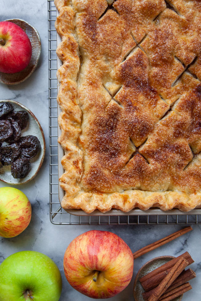 An apple and prunes slab pie next to apples, prunes and sticks of cinnamon