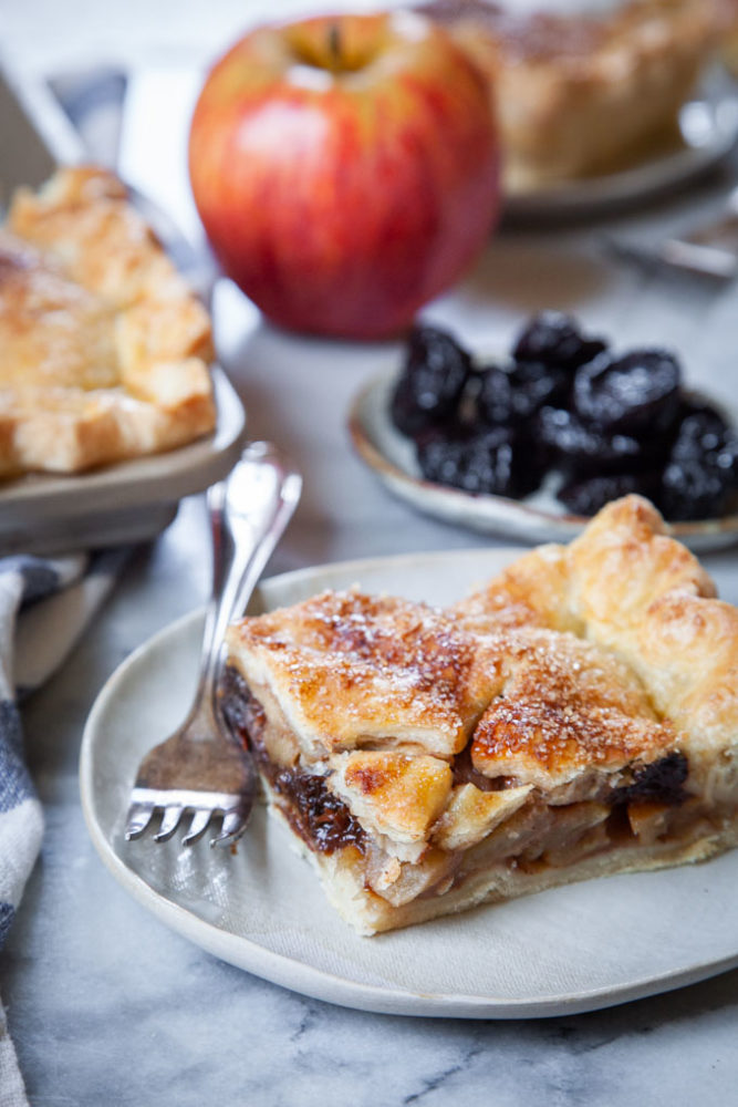 A slice of apple slab pie on a plate, with apples and prunes behind it, along with the remaining pie to the side.