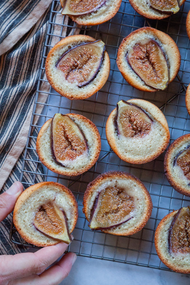 A hand reaching for fig financier tea almond cakes on wire cooling rack.