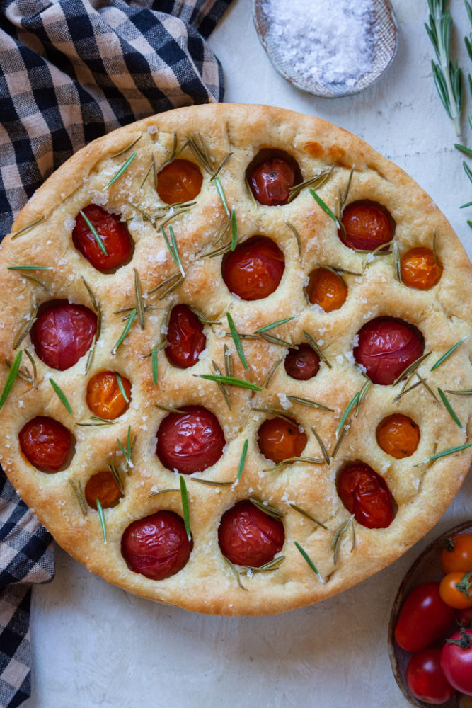 Cherry tomato and rosemary focaccia on a table with some salt, cherry tomatoes and rosemary herbs next to it.